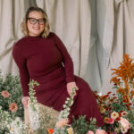 A woman wearing a long burgundy dress sits on a wooden stool surrounded by colorful flowers, smiling toward the camera in front of soft draped fabric.