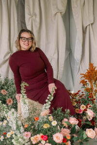 A woman wearing a long burgundy dress sits on a wooden stool surrounded by colorful flowers, smiling toward the camera in front of soft draped fabric.