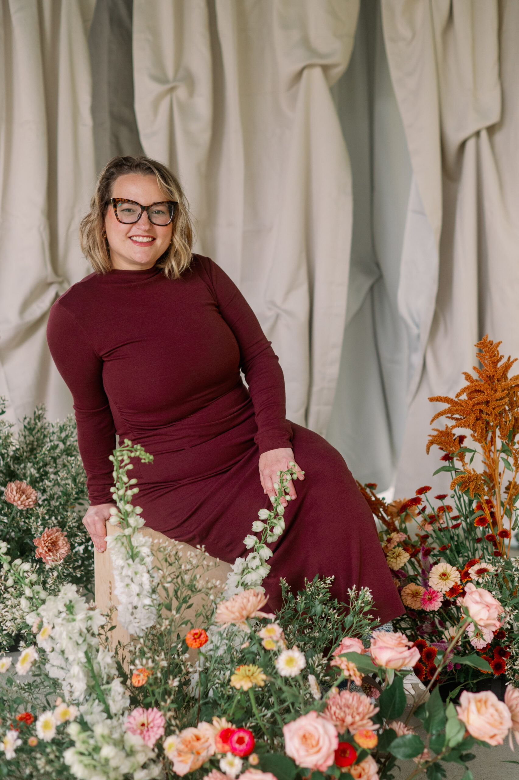 A woman wearing a long burgundy dress sits on a wooden stool surrounded by colorful flowers, smiling toward the camera in front of soft draped fabric.
